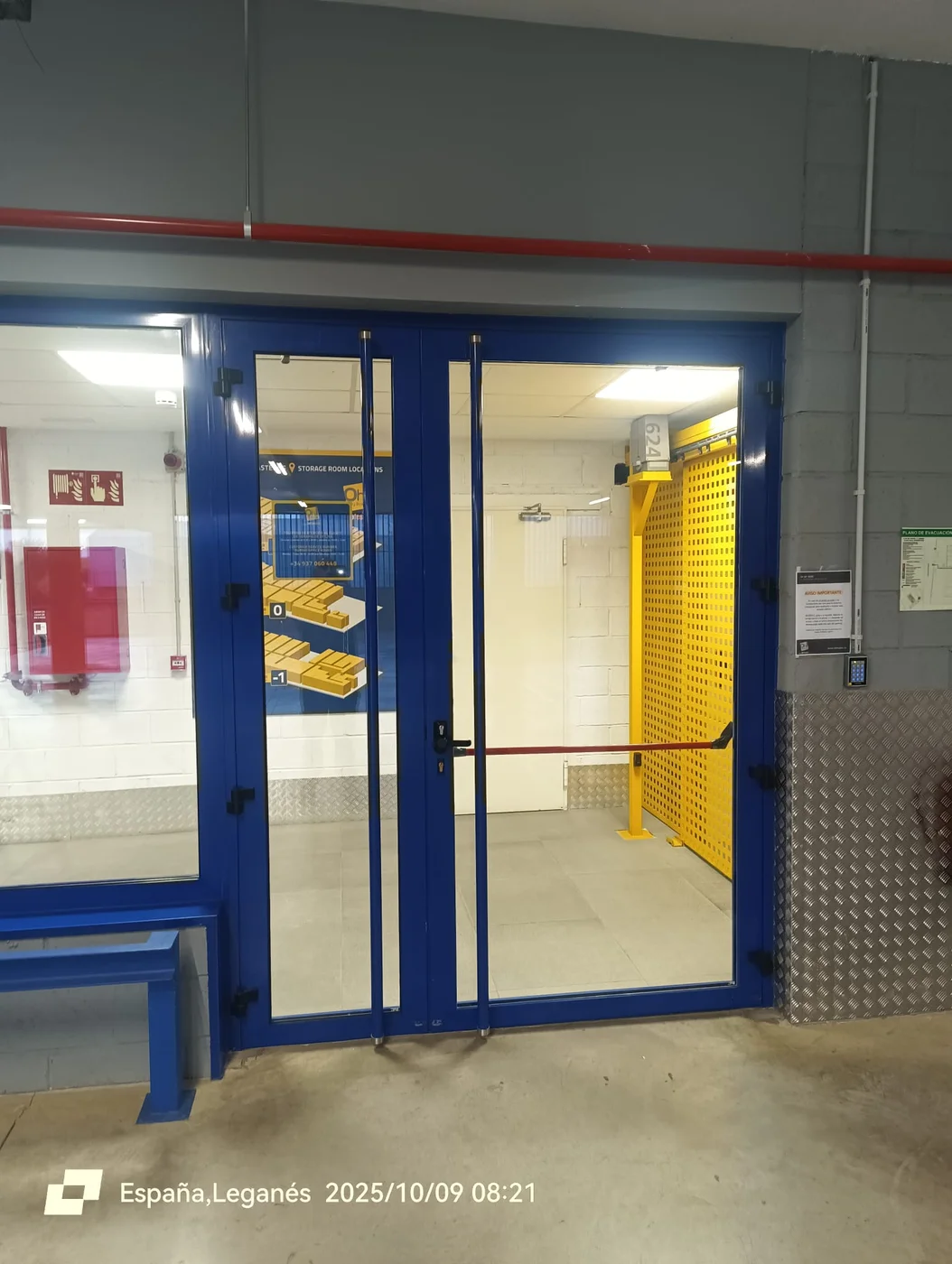 Puerta peatonal de dos hojas acristaladas en azul con tiradores verticales y barra antipánico en Leganés
